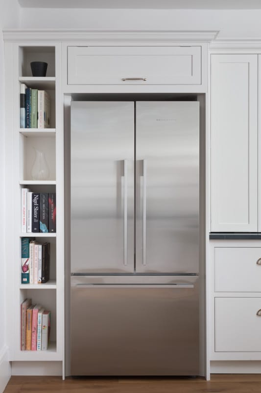 Silver American style refrigerator surrounded by white Arbor kitchen cabinetry and shelving unit