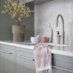 Closeup of sink area featuring green-grey kitchen cabinetry, white worktop, silver sink and tap