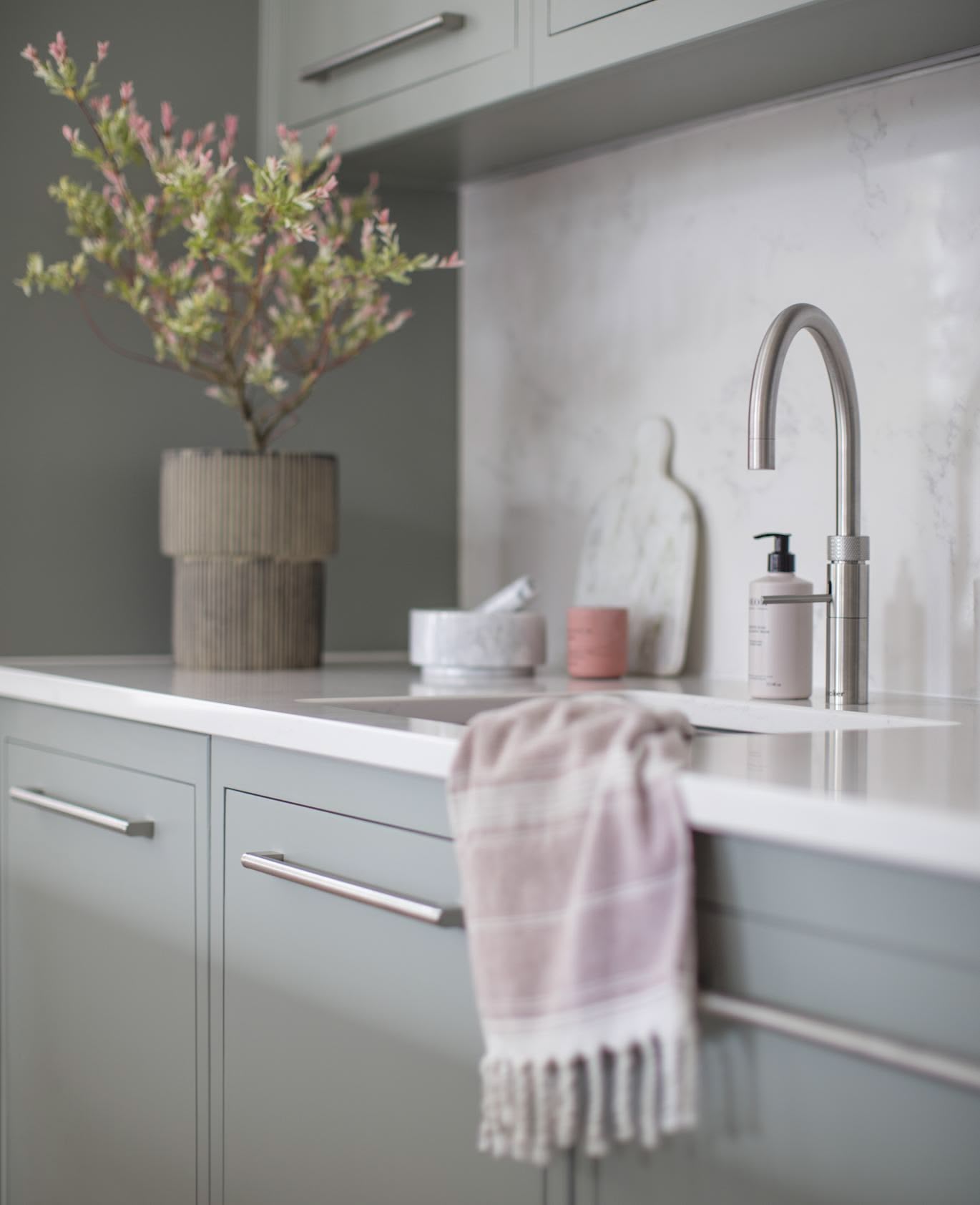 Closeup of sink area featuring green-grey kitchen cabinetry, white worktop, silver sink and tap