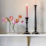 White shelving featuring gold rail detailing holding black candle sticks and potted pink flowers