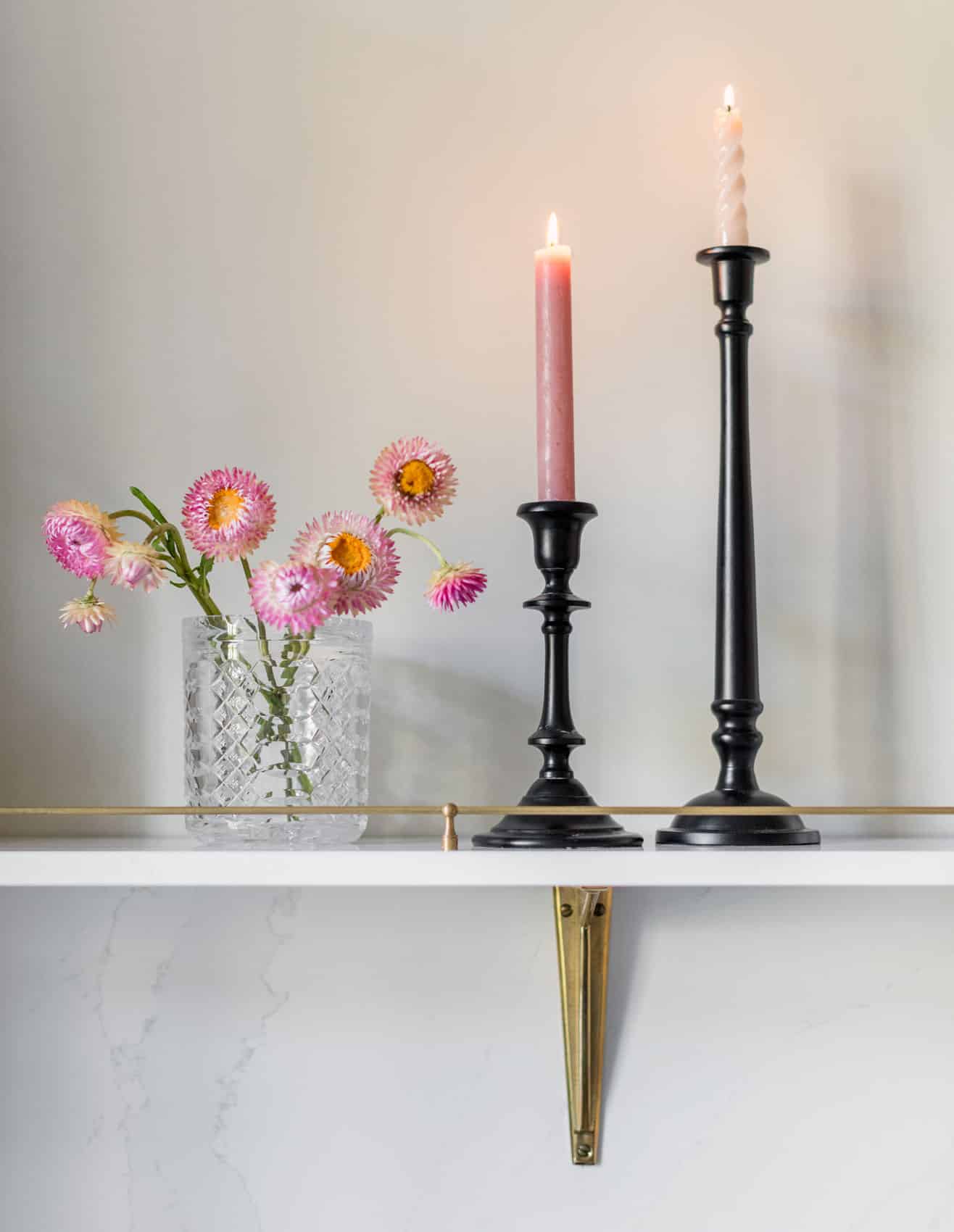 White shelving featuring gold rail detailing holding black candle sticks and potted pink flowers