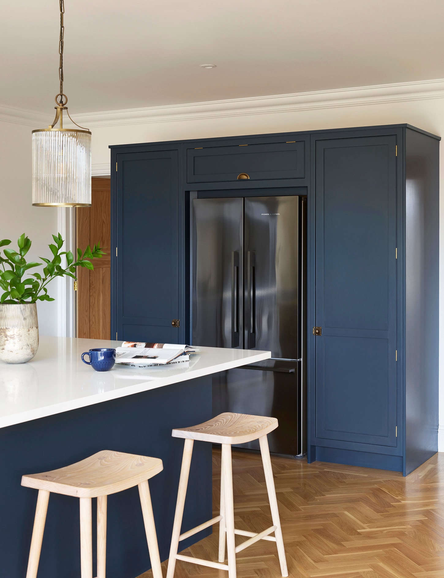 Deep blue kitchen island with white worktop in front of an American style refrigerator surrounded by Arbor cabinetry