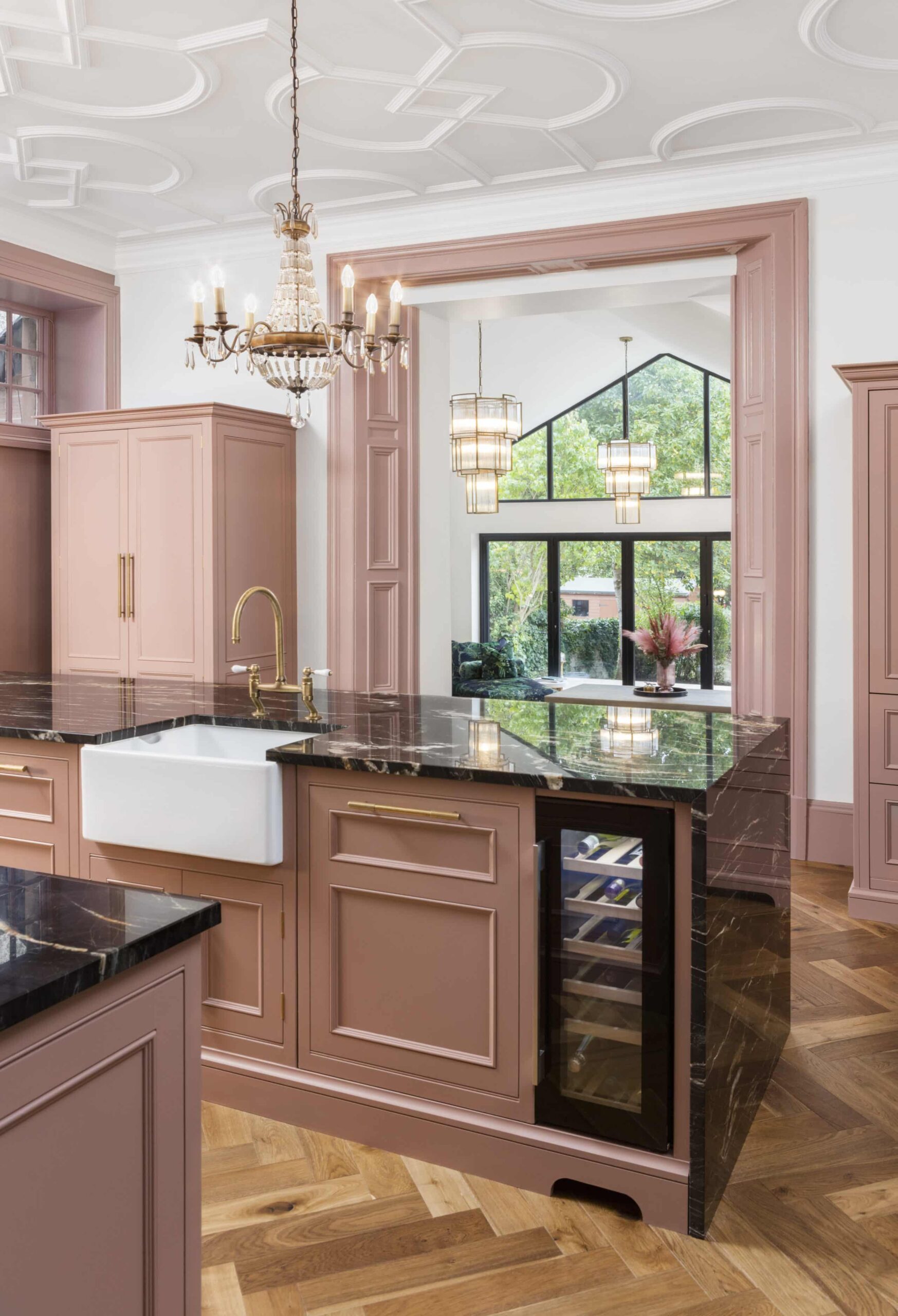 Pink kitchen island with black marble worktop and white sink with gold tap featuring wine fridge