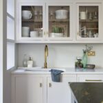 White Linear kitchen cabinetry with glass window doors above white sink with gold tap