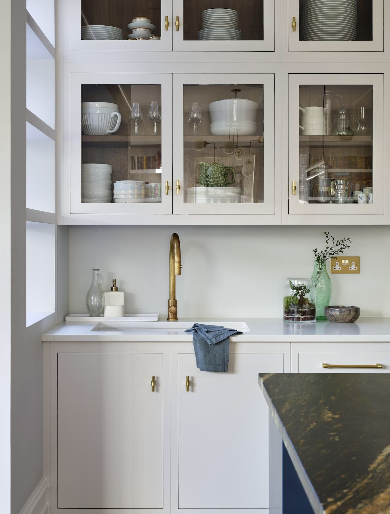 White Linear kitchen cabinetry with glass window doors above white sink with gold tap