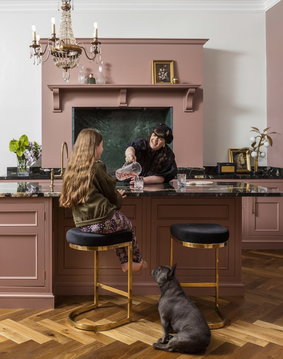 Mother and young daughter around pink kitchen island with black worktop featuring gold and black stools