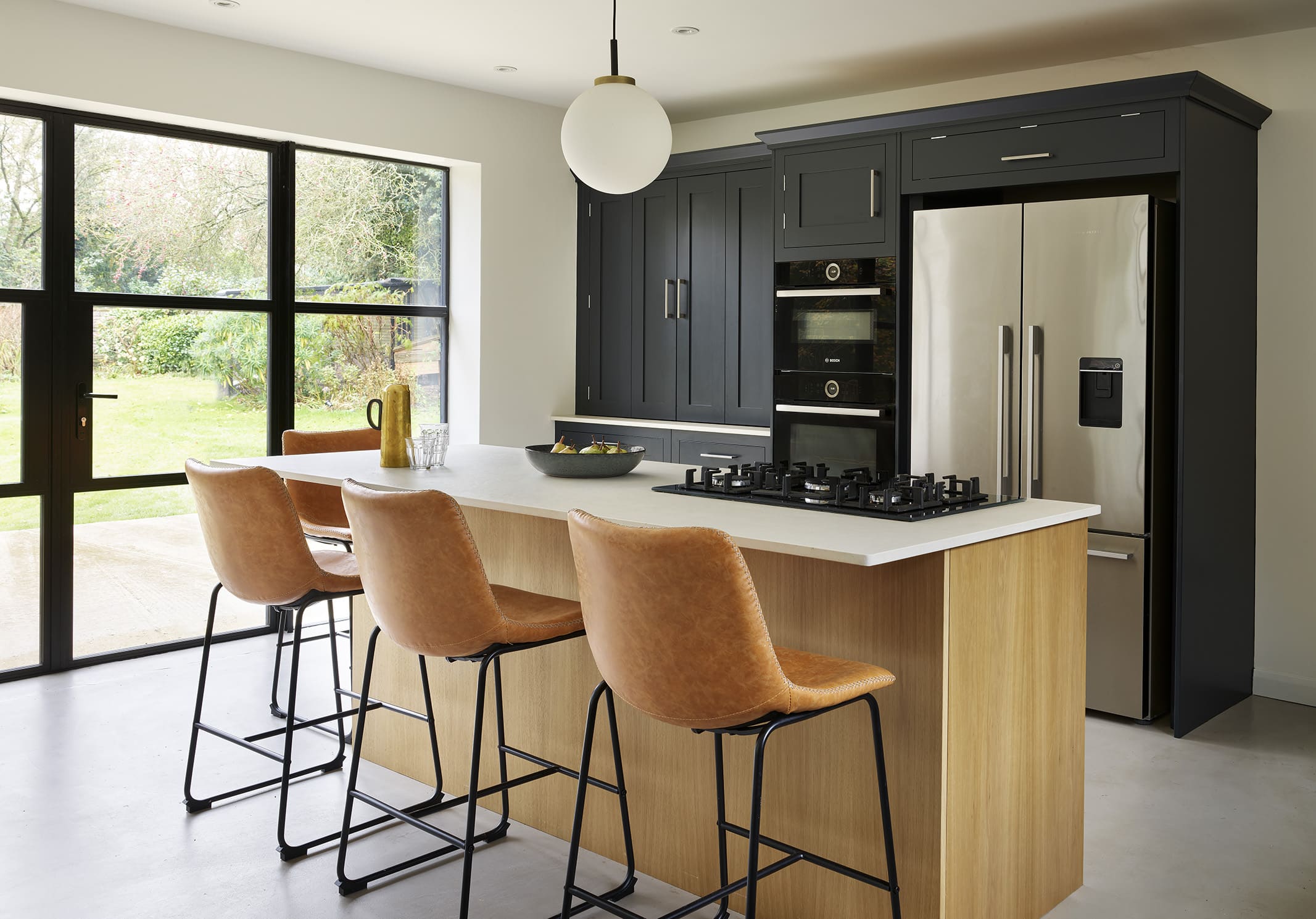 Light oak Shaker kitchen island with white worktop and orange stools overlooking dark Shaker kitchen cabinetry