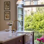 Burgundy kitchen island with white worktop and basin sink featuring a gold tap in front of open French doors