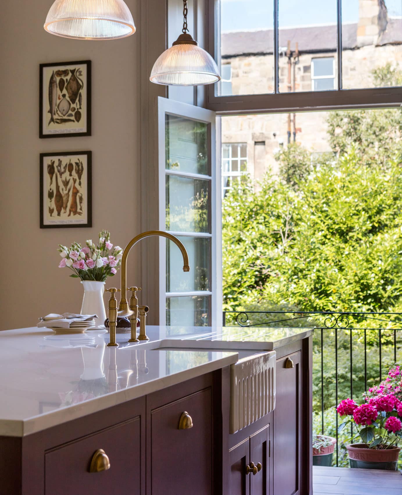 Burgundy kitchen island with white worktop and basin sink featuring a gold tap in front of open French doors