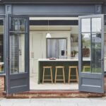Open French doors looking into an Arbor kitchen featuring a green and white kitchen island with wooden stools