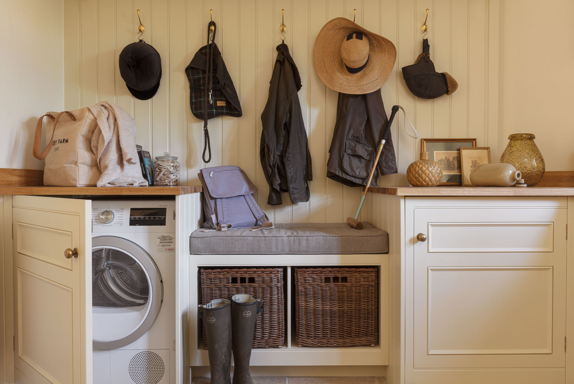 Cream and white boot room featuring grey bench seating, brown storage baskets and coat hooks above
