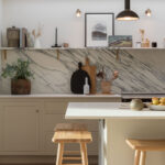 Beige kitchen island with white worktop and wooden stools in front of beige kitchen cabinetry and marble splashback