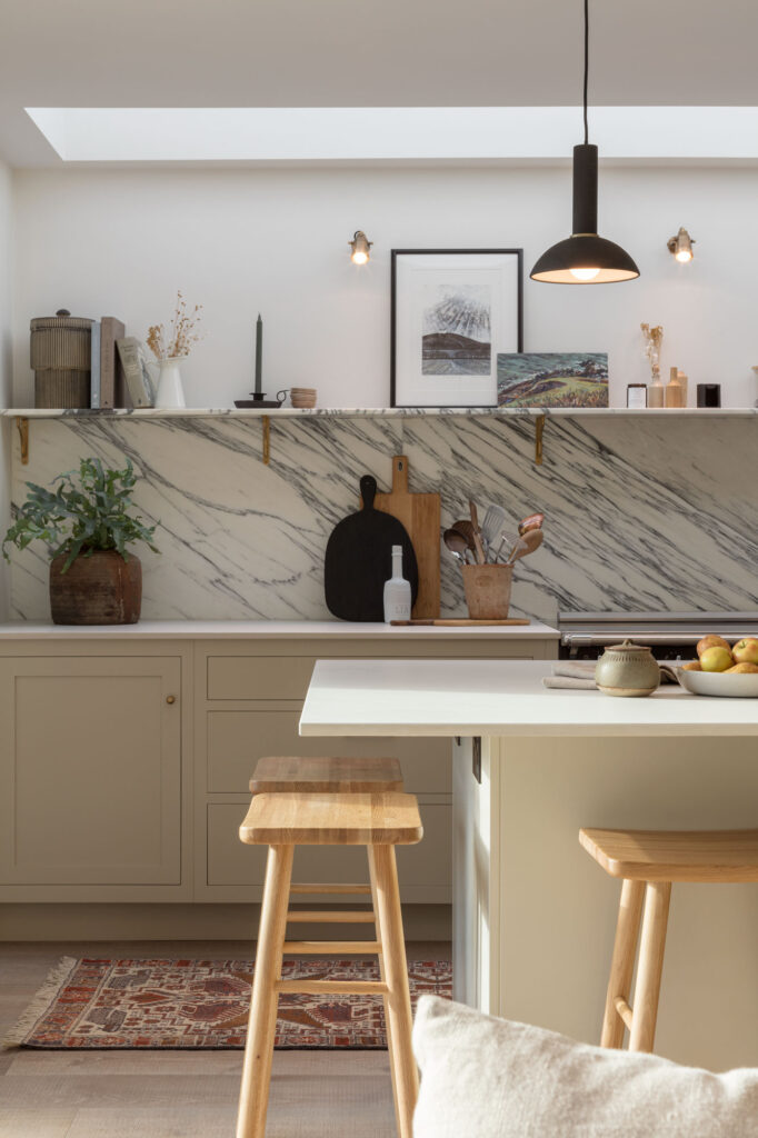Beige kitchen island with white worktop and wooden stools in front of beige kitchen cabinetry and marble splashback