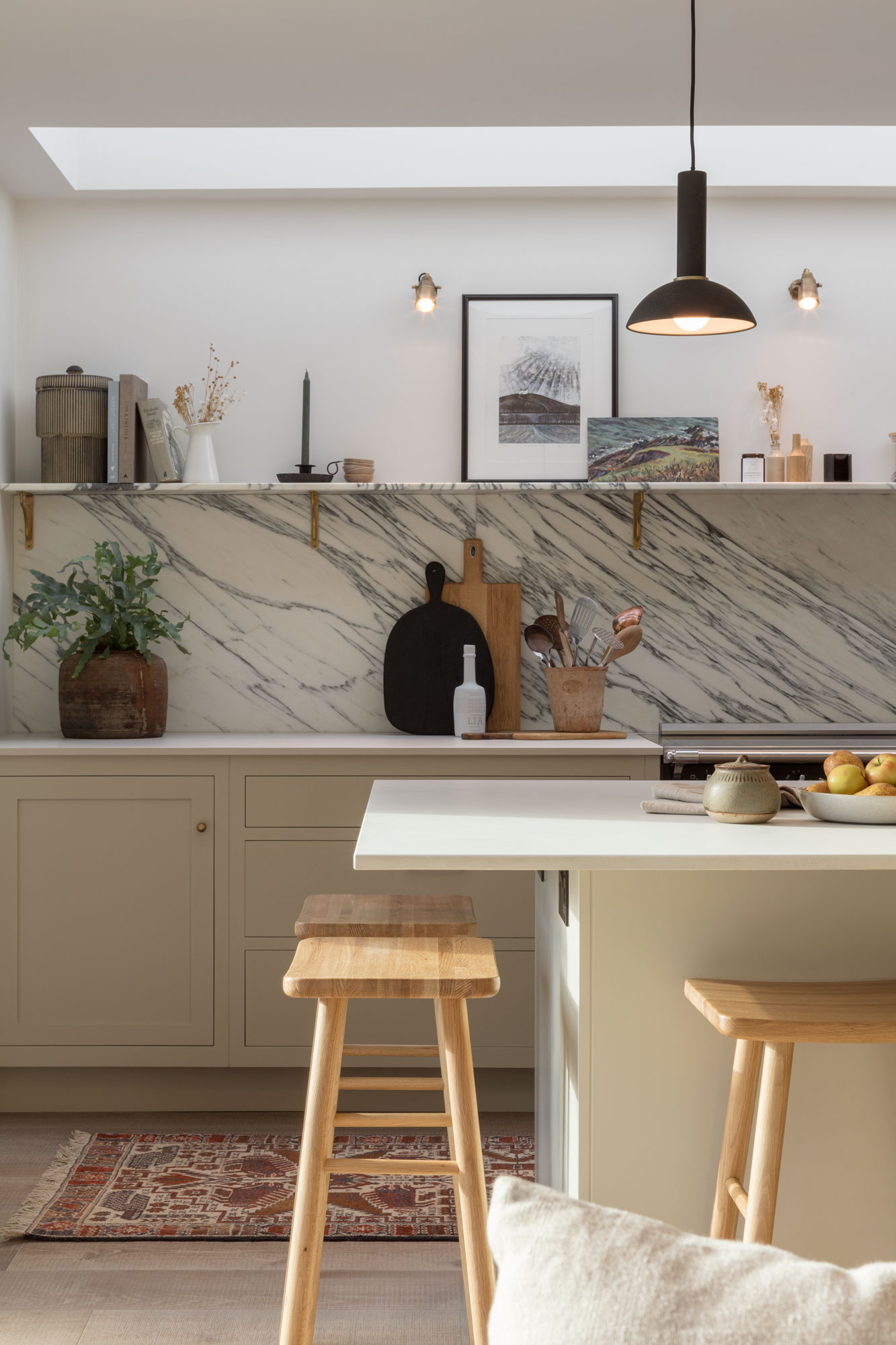 Beige kitchen island with white worktop and wooden stools in front of beige kitchen cabinetry and marble splashback