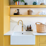 Bespoke utility room with bright yellow cabinetry, ceramic sink, and open shelving with lighting for laundry and cleaning essentials.