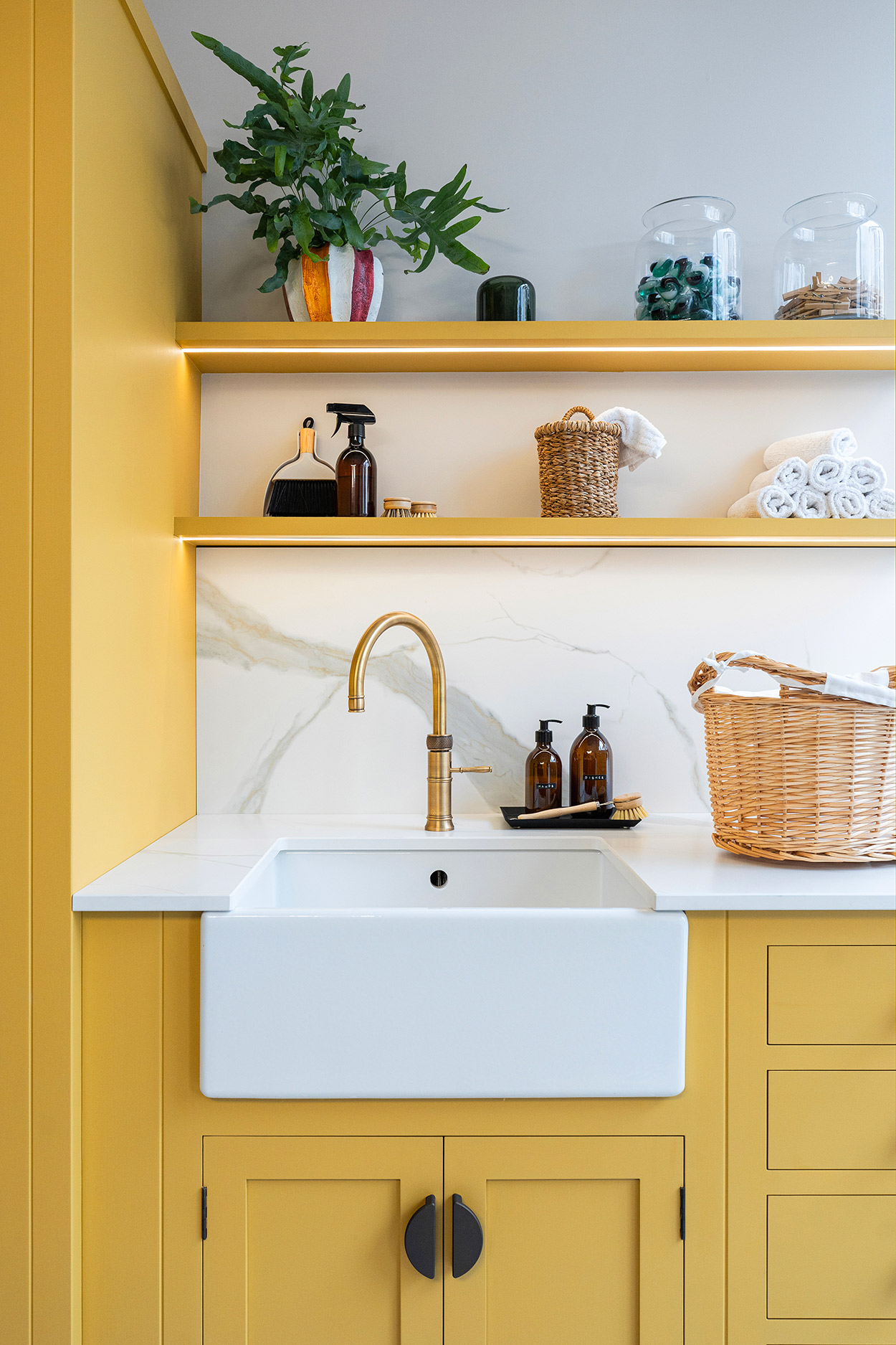 Bespoke utility room with bright yellow cabinetry, ceramic sink, and open shelving with lighting for laundry and cleaning essentials.