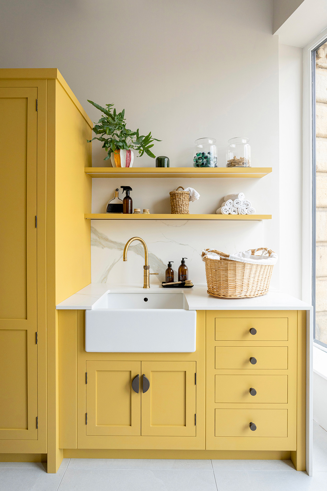 Yellow bespoke utility cabinetry with open shelves, brass tap, white basin, and wicker laundry basket.