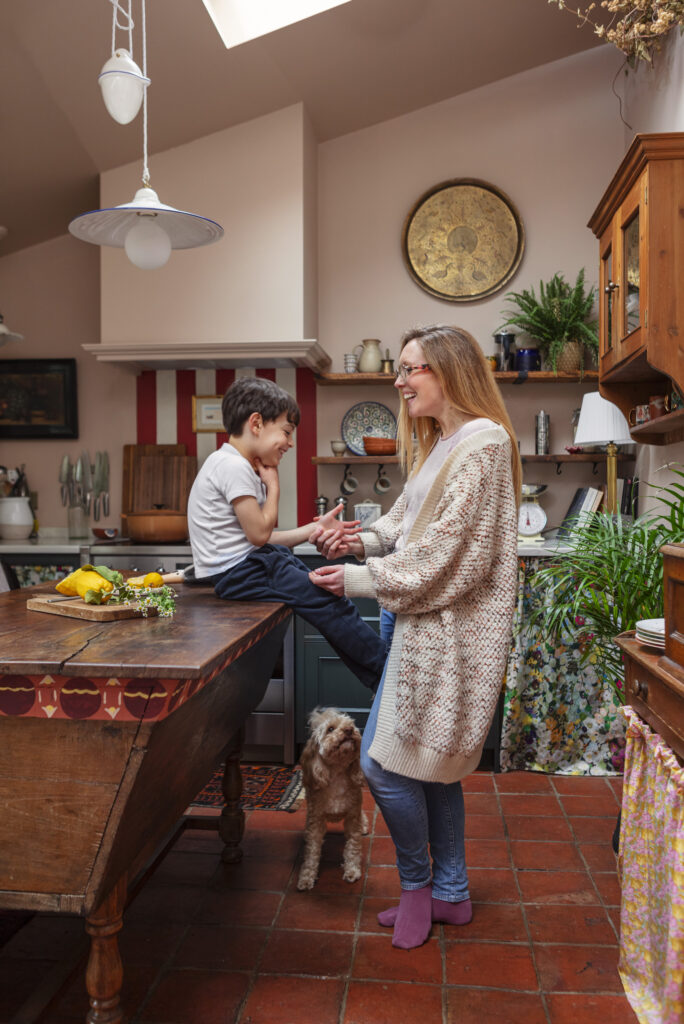 child sitting on table and mother standing laughing in front of kitchen with vibrant wallpaper in the kitchen and heart of the home