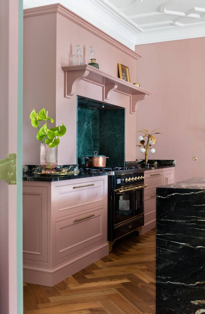 Blush pink hand-painted kitchen with a black range cooker, green marble splashback and herringbone wood flooring.