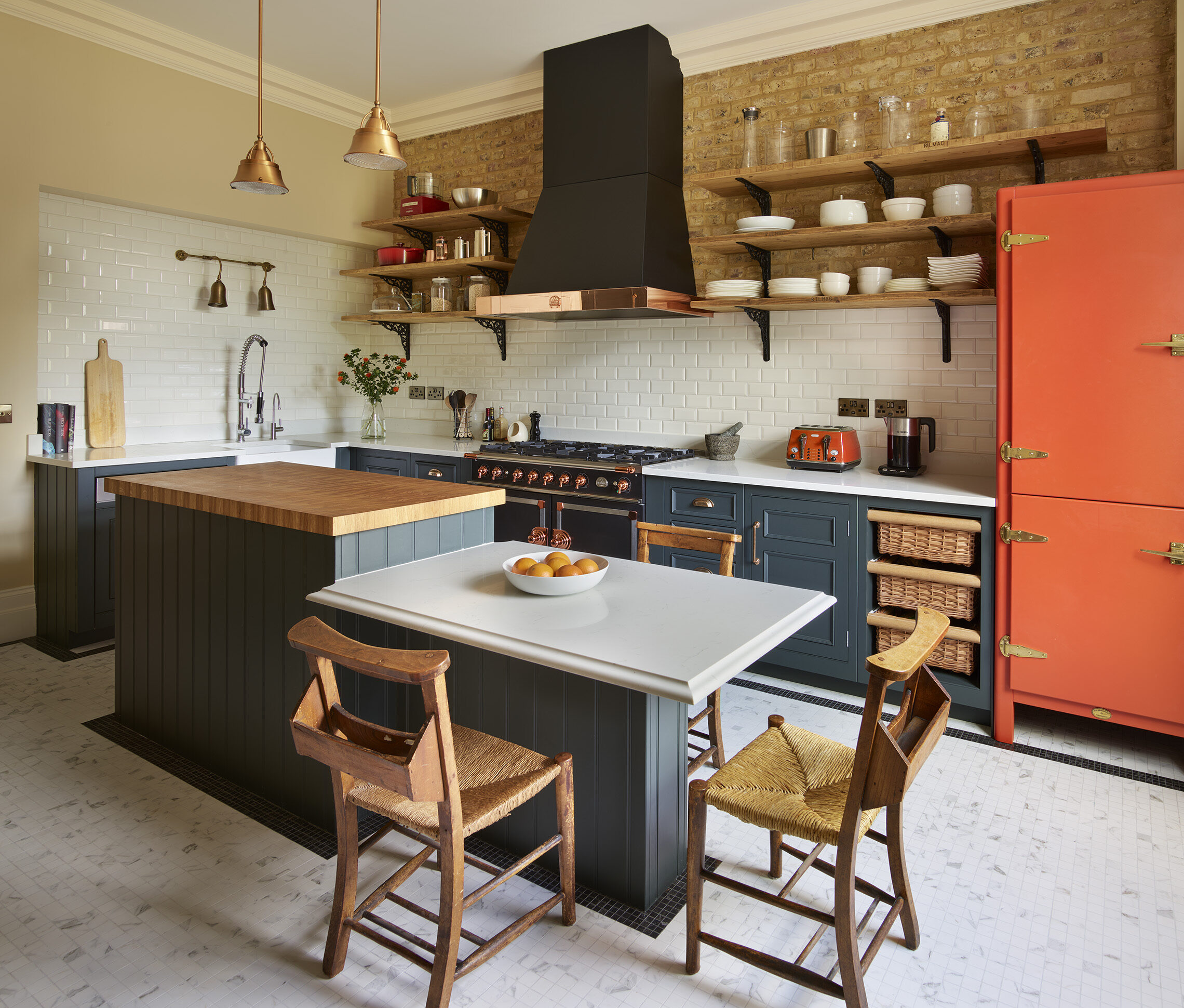 A modern kitchen featuring dark cabinetry, a two-tier central island with a light worktops and wooden stools, and white subway tile splashback. Shelves with various utensils and copper pendant lights complement the space.