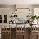 Elegant kitchen interior featuring a marble backsplash, beige cabinetry, and a central island with wooden stools. Decorative plants and bowls of fruit add a welcoming touch.