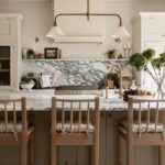 Elegant kitchen interior featuring a marble backsplash, beige cabinetry, and a central island with wooden stools. Decorative plants and bowls of fruit add a welcoming touch.