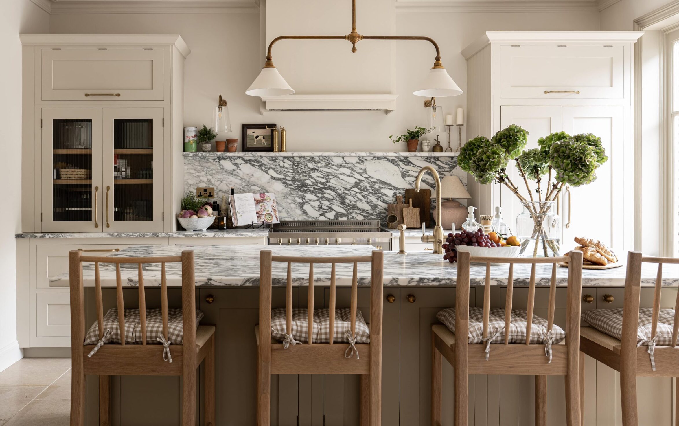 Elegant kitchen interior featuring a marble backsplash, beige cabinetry, and a central island with wooden stools. Decorative plants and bowls of fruit add a welcoming touch.