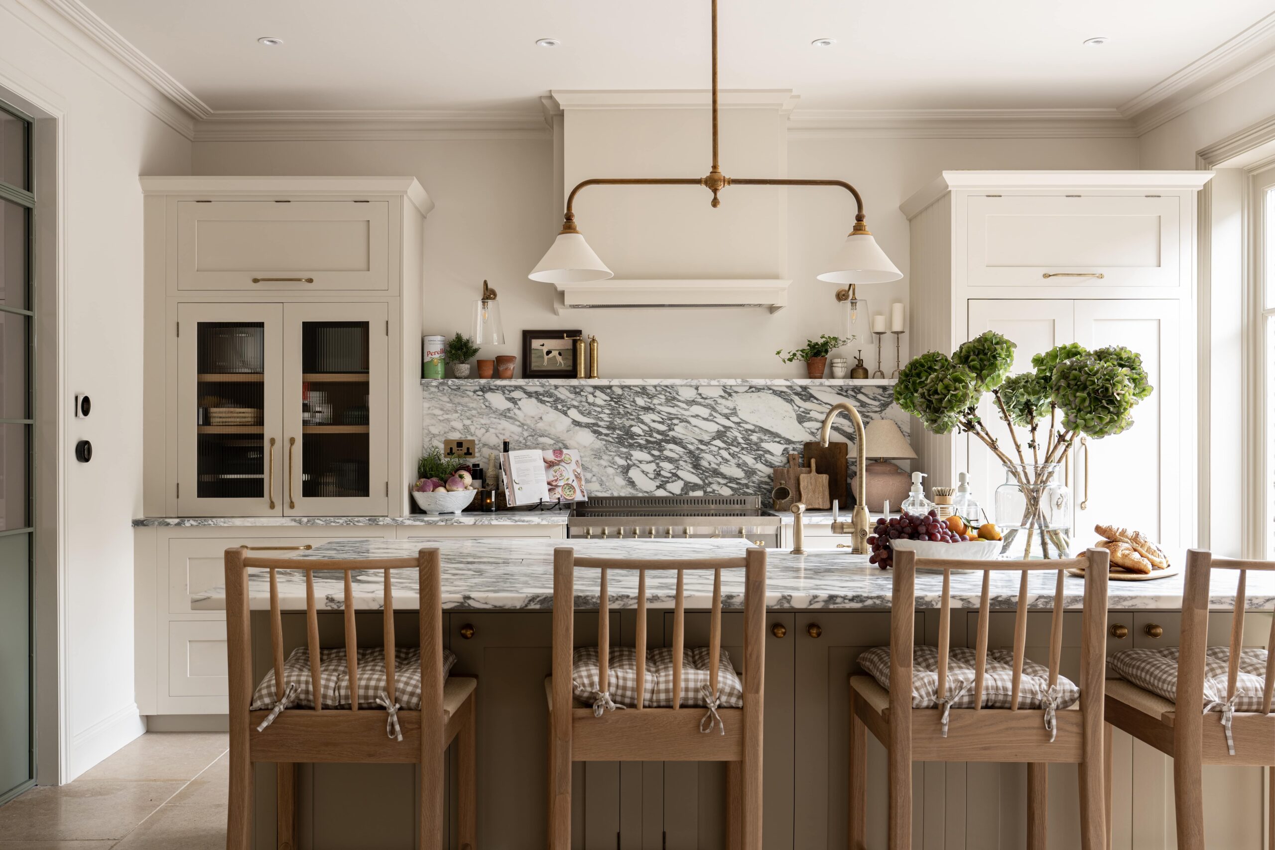 Elegant kitchen interior featuring a marble backsplash, beige cabinetry, and a central island with wooden stools. Decorative plants and bowls of fruit add a welcoming touch.