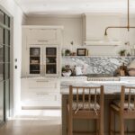 A vertical image of a modern kitchen featuring white cabinetry, a marble countertop, and stainless steel appliances. A wooden dining table with chairs is in the foreground. Natural light streams in through a glass door on the left.