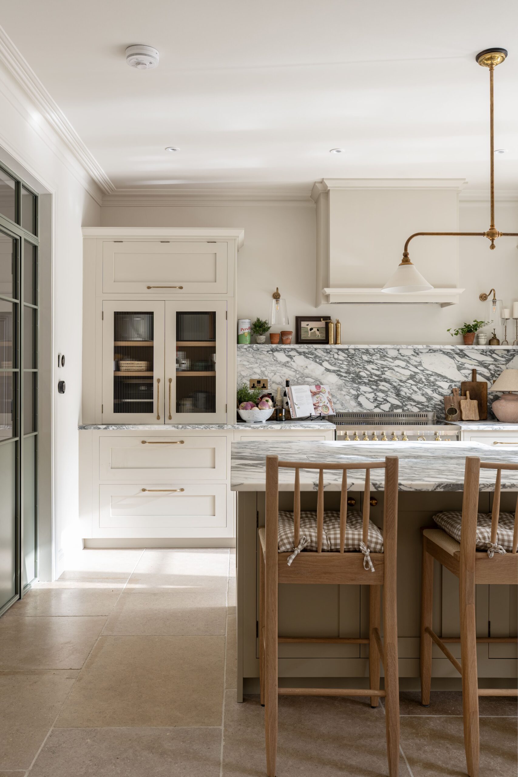 A vertical image of a modern kitchen featuring white cabinetry, a marble countertop, and stainless steel appliances. A wooden dining table with chairs is in the foreground. Natural light streams in through a glass door on the left.