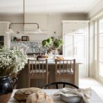 Elegant kitchen interior with marble backsplash, warm neutral cabinetry, and a wooden dining table set with fruits and plants.