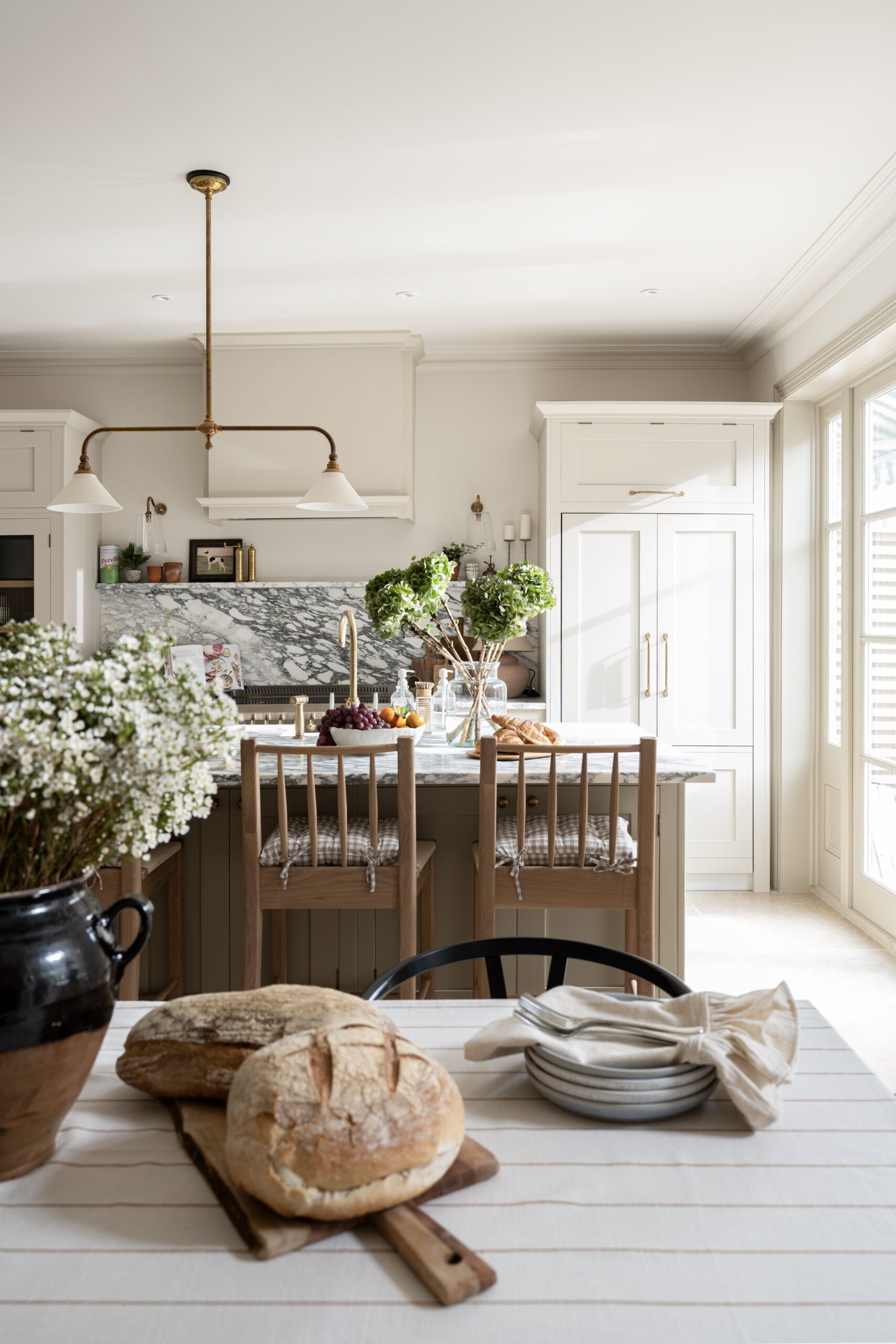 Elegant kitchen interior with marble backsplash, warm neutral cabinetry, and a wooden dining table set with fruits and plants.