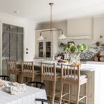 Interior of a spacious kitchen featuring a central island with stools, and a dining area with a wooden table set with food items. The design includes white cabinetry and modern appliances. Natural light streams in through large windows.