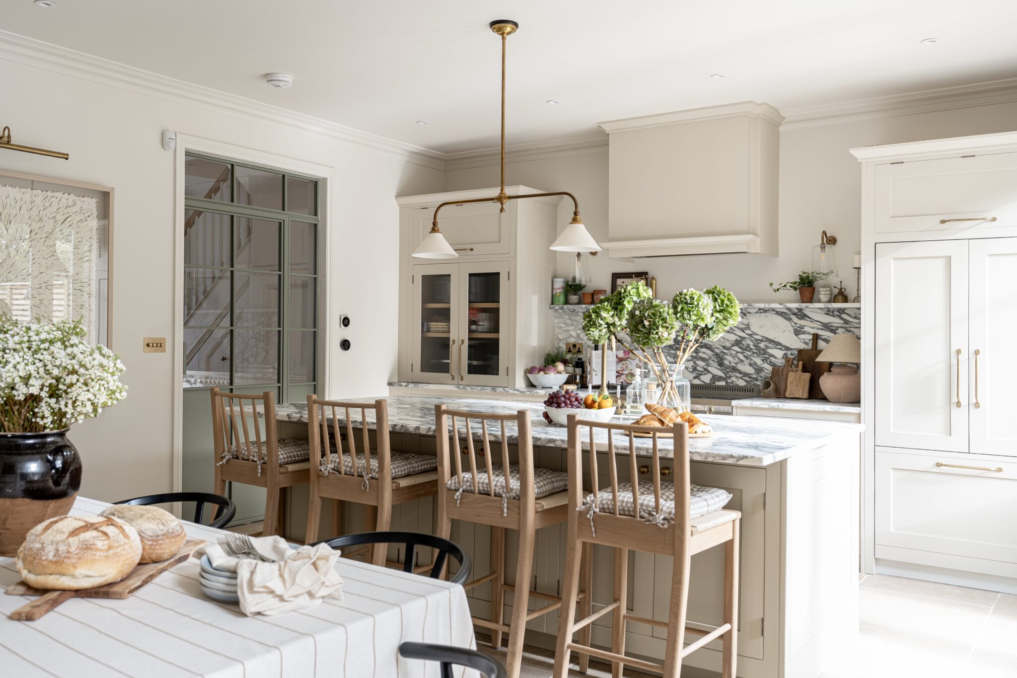 Interior of a spacious kitchen featuring a central island with stools, and a dining area with a wooden table set with food items. The design includes white cabinetry and modern appliances. Natural light streams in through large windows.