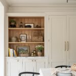 Interior of a tidy kitchen dining area featuring beige cabinetry, wooden shelving decorated with books and plants, and a small dining area with a striped tablecloth and a black chair.