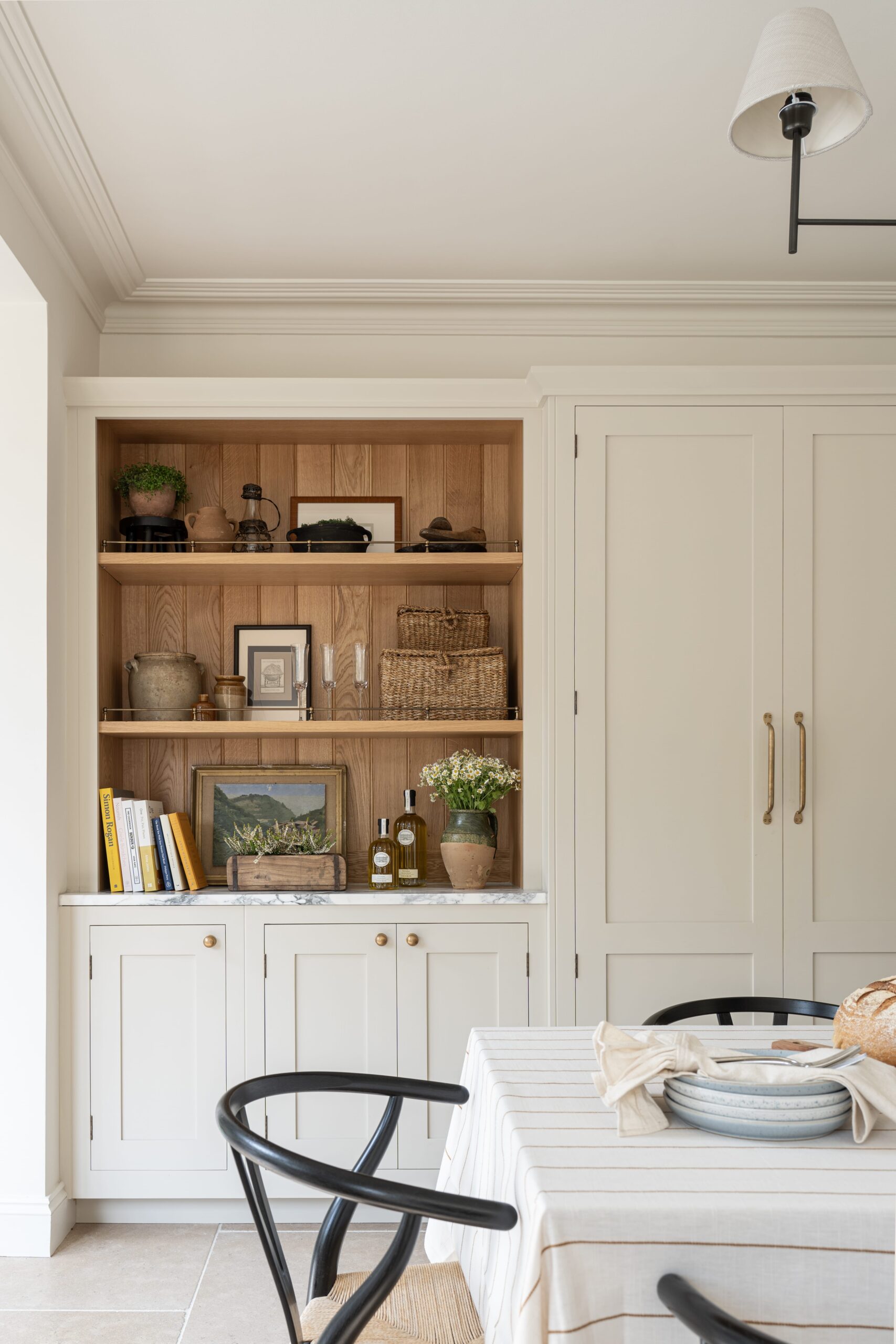 Interior of a tidy kitchen dining area featuring beige cabinetry, wooden shelving decorated with books and plants, and a small dining area with a striped tablecloth and a black chair.