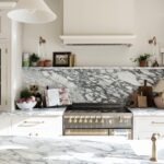 Modern kitchen with marble countertops and backsplashes, featuring white cabinetry and equipped with high-end appliances. Decorative plants and cooking utensils are neatly arranged, adding to the overall elegance of the space.