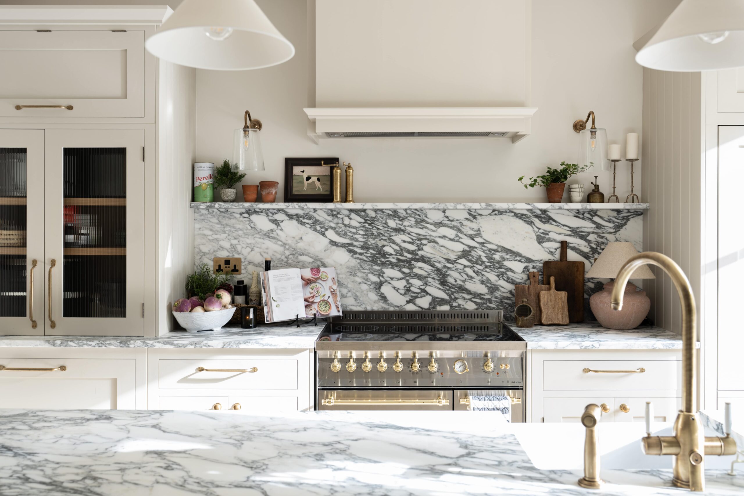 Modern kitchen with marble countertops and backsplashes, featuring white cabinetry and equipped with high-end appliances. Decorative plants and cooking utensils are neatly arranged, adding to the overall elegance of the space.