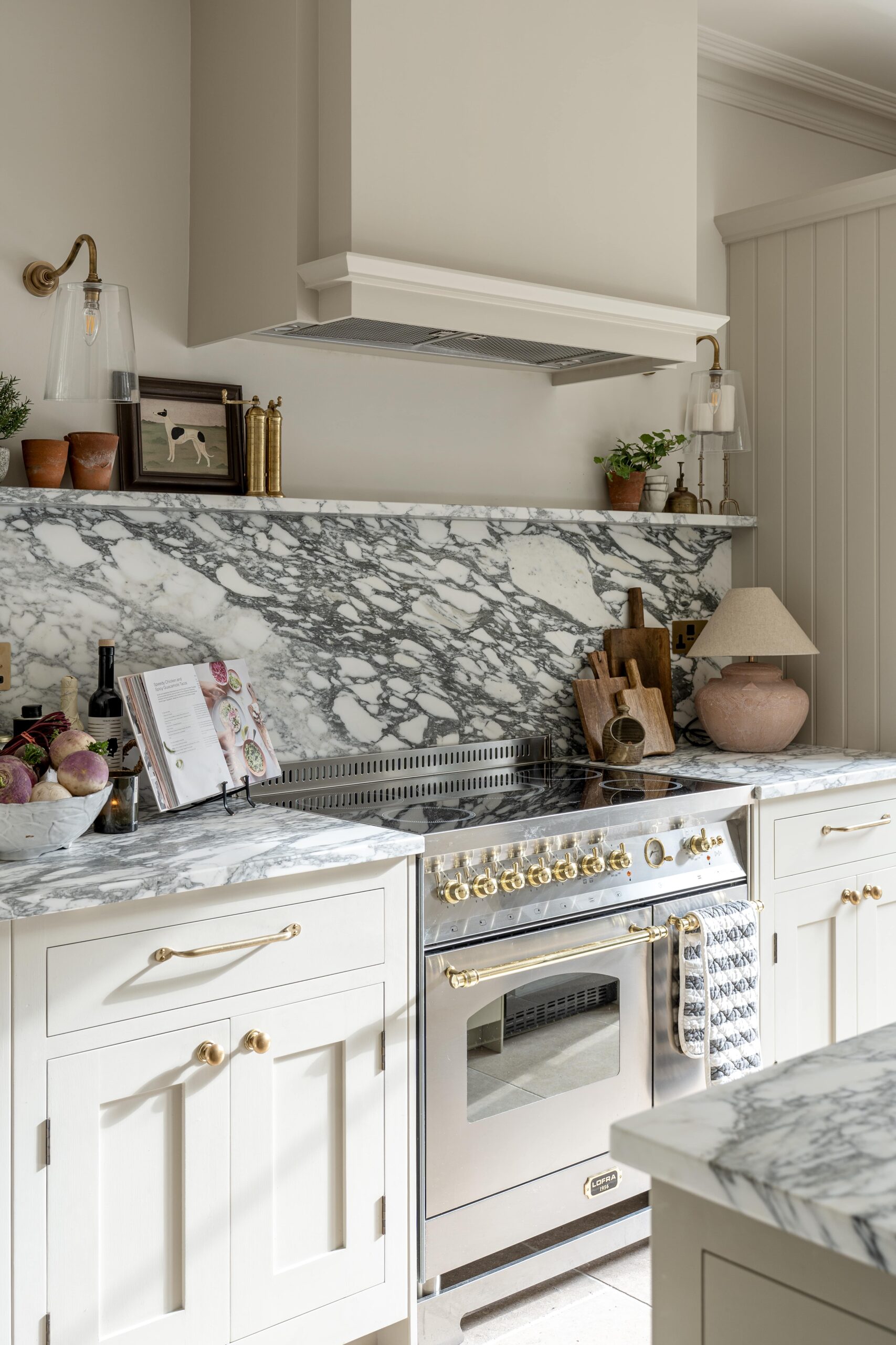 Elegant kitchen featuring a marble backsplash and countertops, with a white cabinetry and a La Cornue range oven. Decor includes brass accents and neatly arranged cooking utensils.