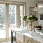 Bright kitchen featuring marble countertops, white cabinetry, and a large window with a view of greenery. Decor includes fresh flowers and assorted kitchen items.