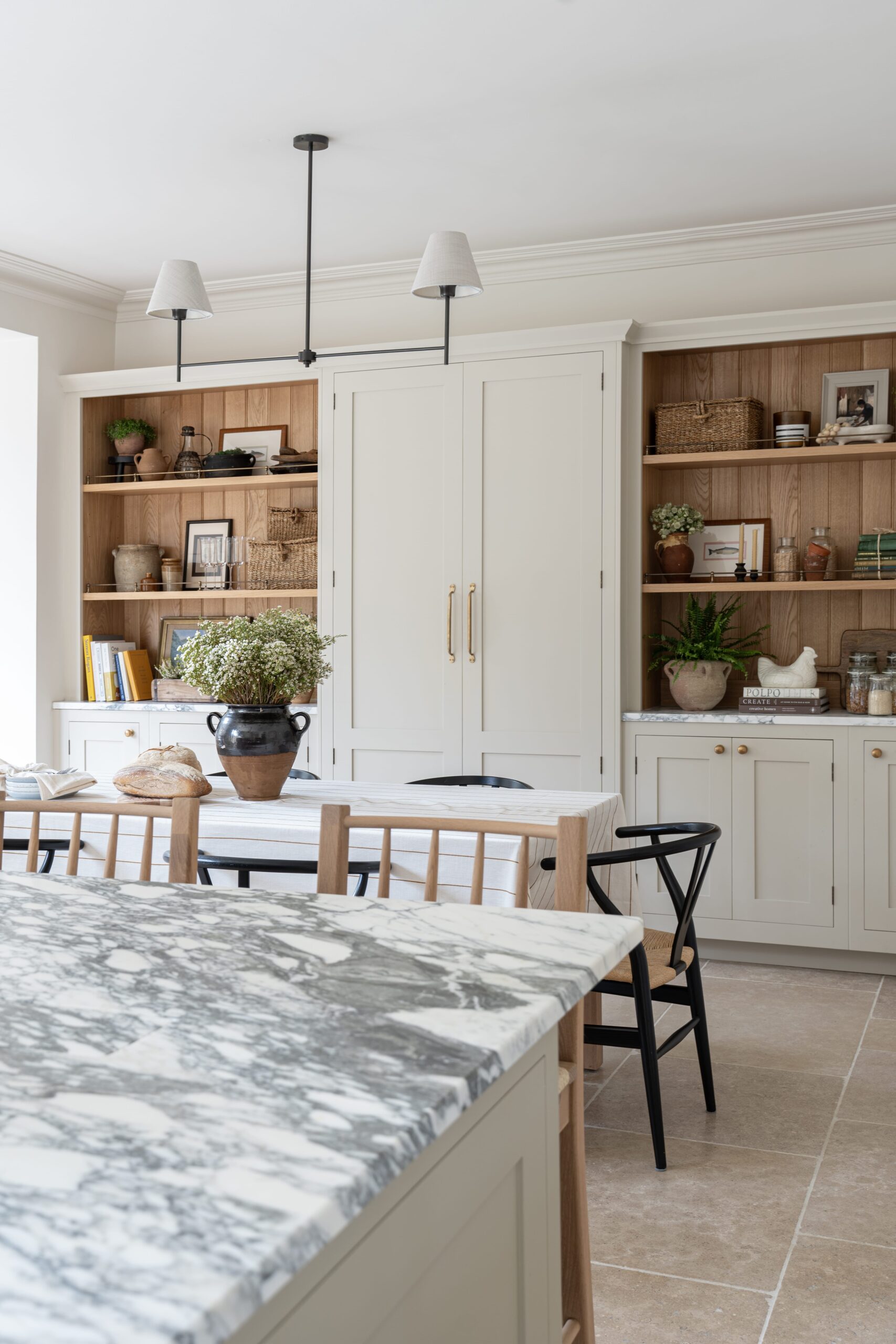 Interior view of a modern kitchen featuring cream cabinets and wooden shelves stocked with various items. In the foreground is a marble island worktop with wooden stools. Black chairs are placed at the dining table, and pendant lights hang above.