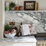 Modern kitchen countertop featuring a marble surface with a basket of fresh vegetables, cooking books propped open, and decorative items.