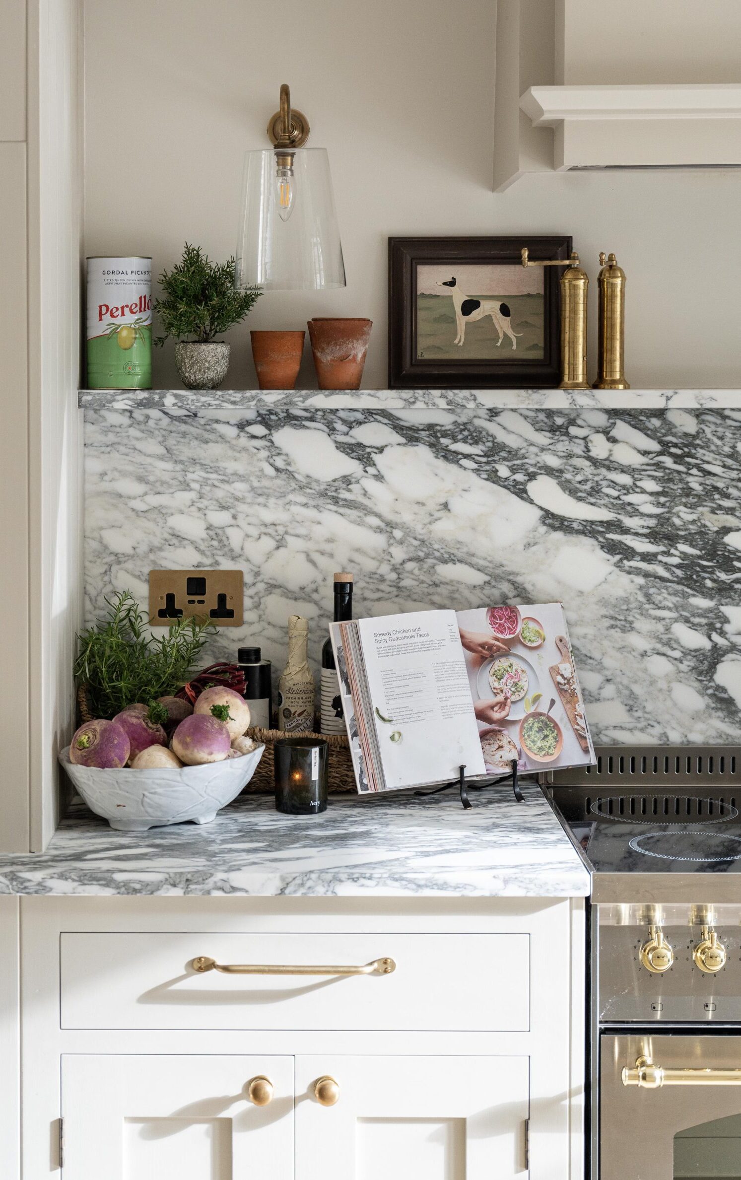 Modern kitchen countertop featuring a marble surface with a basket of fresh vegetables, cooking books propped open, and decorative items.