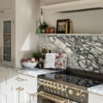 Elegant kitchen interior featuring white cabinetry, a marble backsplash, and gold-colored fixtures. A bowl of fruit, decorative items, and a cookbook are arranged on the counter.