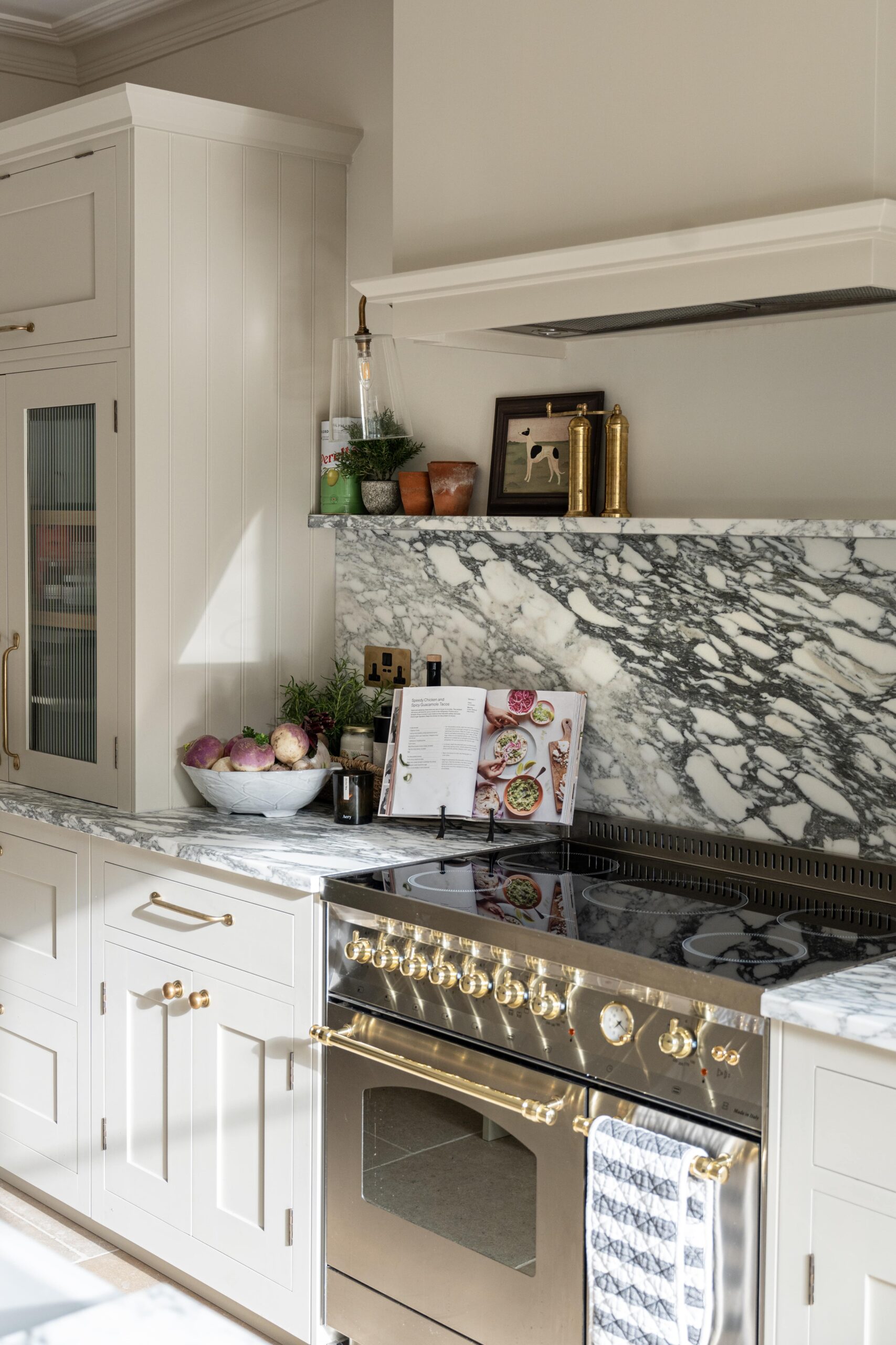 Elegant kitchen interior featuring white cabinetry, a marble backsplash, and gold-colored fixtures. A bowl of fruit, decorative items, and a cookbook are arranged on the counter.