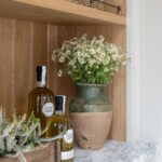 A cosy kitchen shelf displaying two bottles of olive oil, a large ceramic vase with fresh daisies, and a small wooden crate holding glassware. The setting features warm wood tones against a white countertop.