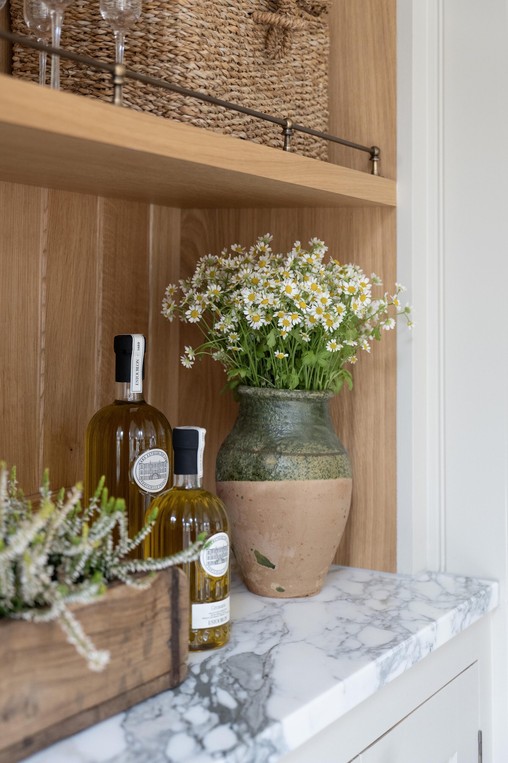 A cosy kitchen shelf displaying two bottles of olive oil, a large ceramic vase with fresh daisies, and a small wooden crate holding glassware. The setting features warm wood tones against a white countertop.