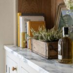 A cosy kitchen shelf displaying a collection of books, including "One Hundred Years of Solitude" by Gabriel Garcia Marquez, a wooden box with blooming heather, and a bottle of olive oil on a marble countertop.