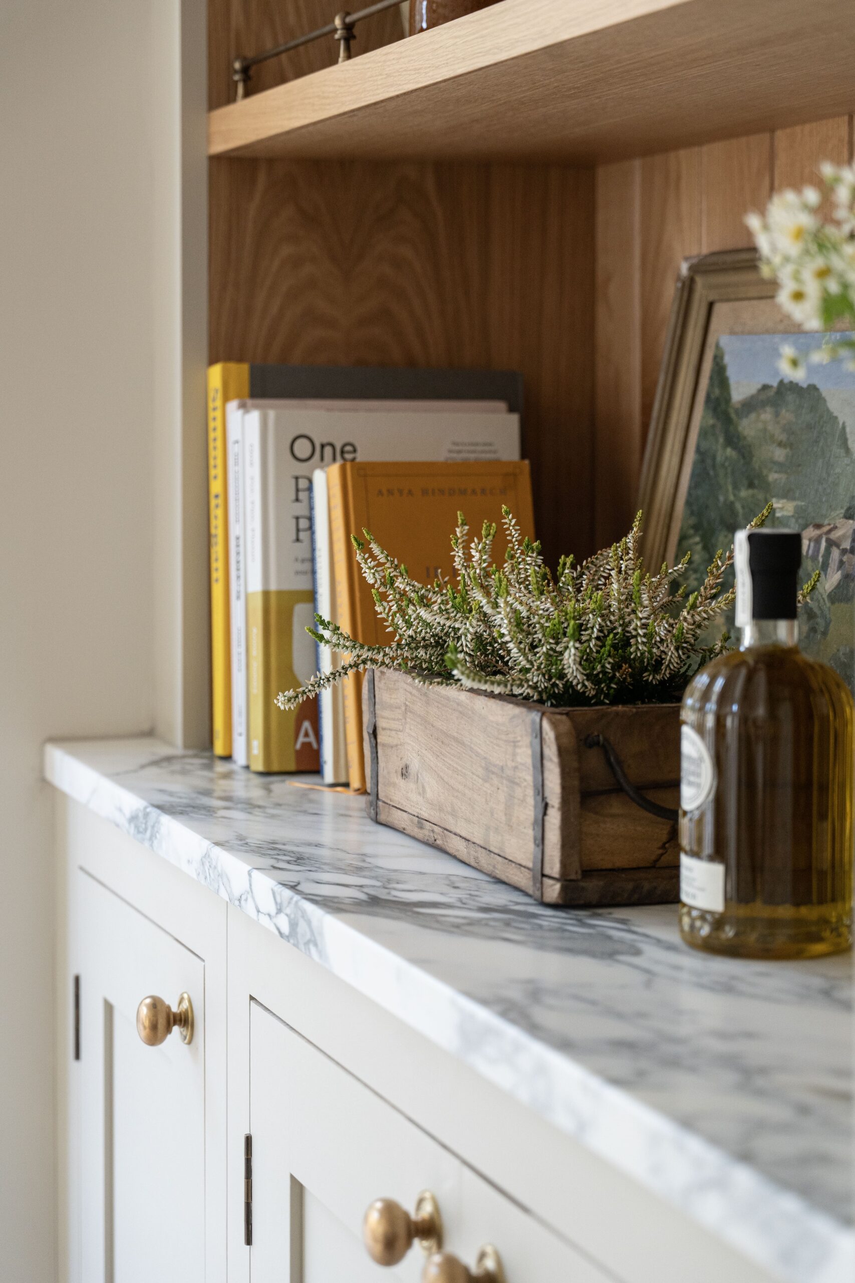 A cosy kitchen shelf displaying a collection of books, including "One Hundred Years of Solitude" by Gabriel Garcia Marquez, a wooden box with blooming heather, and a bottle of olive oil on a marble countertop.
