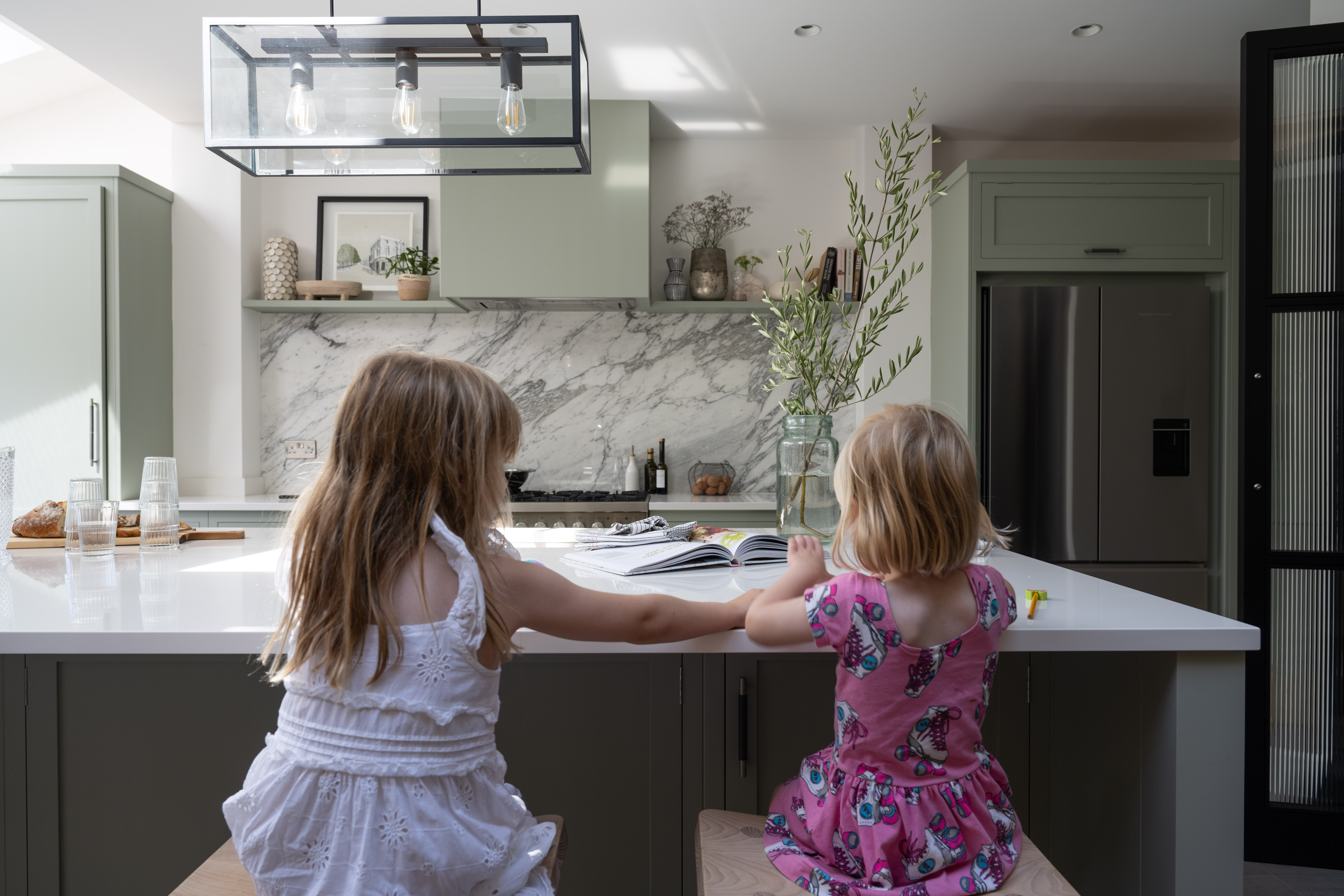 Two children are sitting at a kitchen counter, engaged in drawing. The luxury green kitchen features modern appliances and marble countertops.